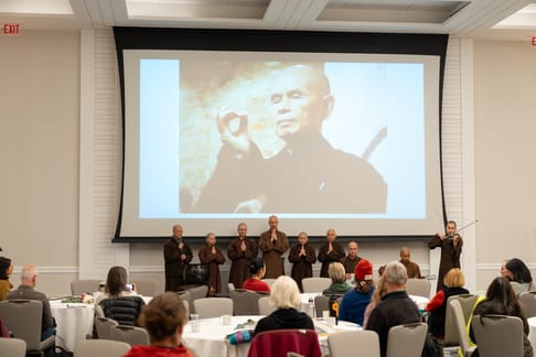 Monastics with prayer hands in front of Thich Nhat Hanh's photo projected on large screen.